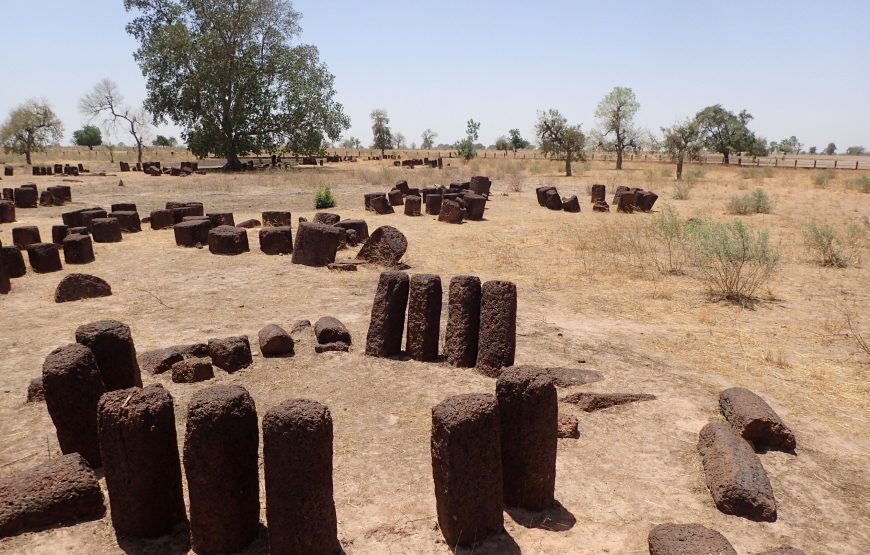 Alt text: Aerial view of megalithic stone circles, an ancient monument of West Africa's civilization, comprising standing stones forming circles with surrounding burial mounds, set against a lush green landscape